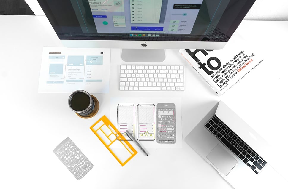 Top down view of a desk with a Mac computer monitor, keyboard, and laptop; a cup of coffee; mobile app wireframes; a book and pen.