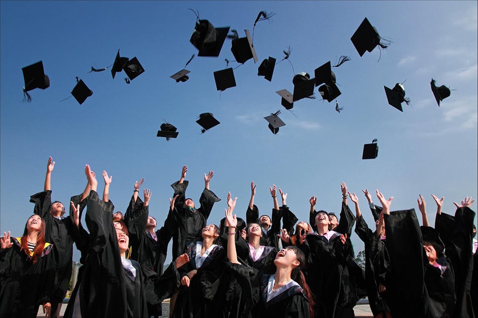 Smiling class of graduates wearing caps and gowns throwing their caps in the air.