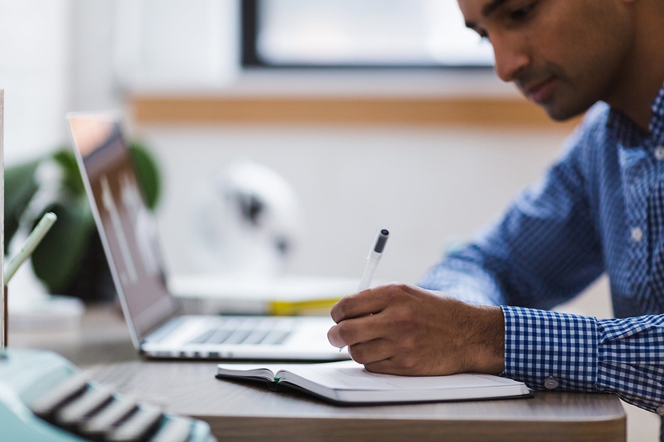 A man sitting at a table with a laptop writing in a notebook.