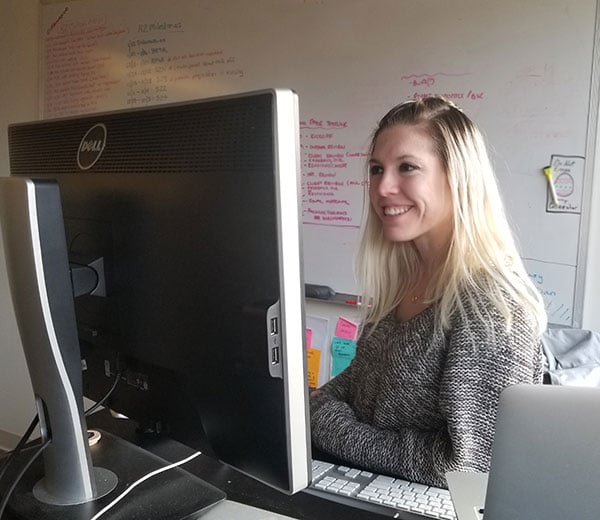 Woman sitting in front of a computer monitor with a white board in the background.