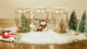 Text that reads "Season's Greetings" in the foreground in white; an old red and white pickup truck, snow on the ground, pine trees, and jars with a tree, snowman, and pine cone in the foreground