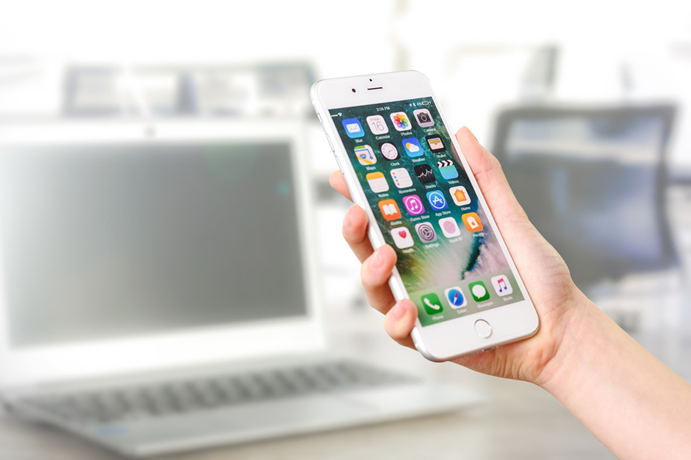 A person's hand holding an iPhone that has lots of apps displayed on the screen. A laptop and office setting are faded in the background.