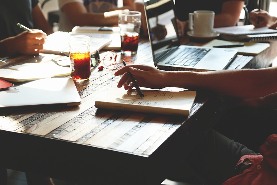 People sitting at a table with a laptop, notebooks, books, and cups of coffee and tea.