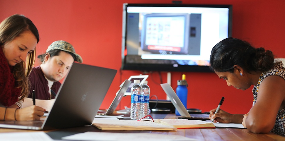 Three people at a table with computers and paper working on a project with a large screen on the wall behind them