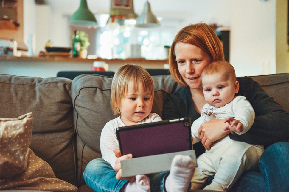 Adult woman sitting on a sofa with two young children playing with an electronic tablet.