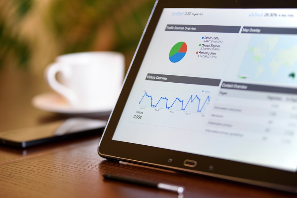 A tablet device displaying a mobile analytics page on a table with a smartphone and coffee cup in the background.